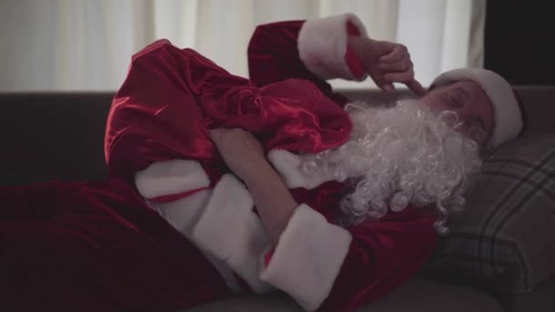 Drunk old man with false white beard in the costume of Santa Claus sleeping on the sofa at home hugging his bag with presents. Bad Santa. Alcoholism, negativism. Camera moving from left to right. — Stock Video