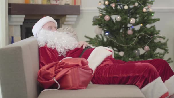 Gammal vit man i jultomte kostym sova på soffan i bakgrunden av Xmas träd. En utmattad kille vilar efter en jobbig arbetsdag hemma. — Stockvideo