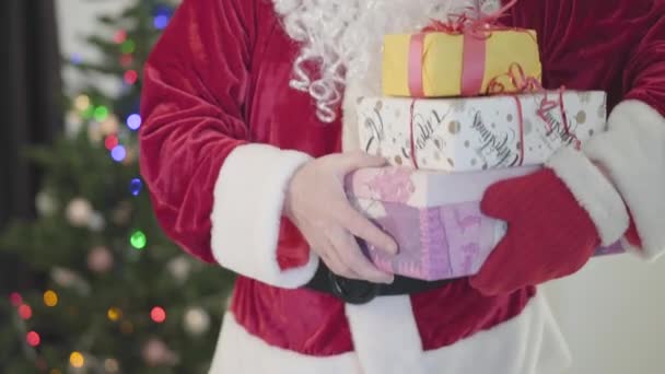 Oude blanke man in kerstman kostuum geeft cadeautjes aan de camera voor de nieuwjaarsboom. Een man met geschenkdozen. Concept van een gelukkige vakantie, tradities, Kerstmis. — Stockvideo