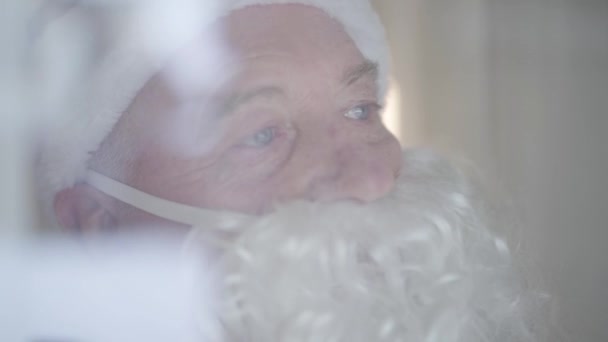 Retrato de cerca del anciano con barba falsa disfrazado de Santa Claus agitando la mano. Concepto de felices fiestas, tradiciones, Navidad. Disparos detrás del vidrio — Vídeos de Stock