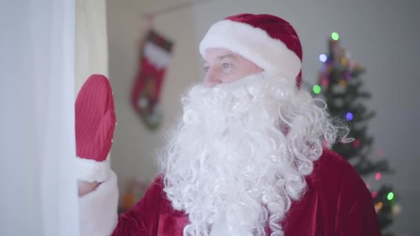 Bătrânul caucazian cu barbă falsă în costum de Moş Crăciun privind pe fereastră şi fluturând mâna. Conceptul de sărbători fericite, tradiții, Crăciun . — Videoclip de stoc