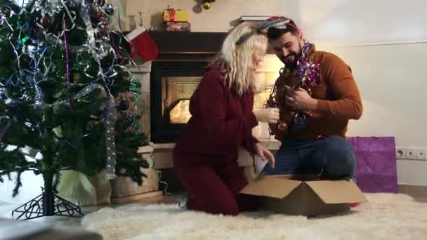 Vidám fehér szőke nő és szakállas férfi ül a kandalló és a karácsonyfa előtt és ukulelézik. Pozitív pár, akik jól érzik magukat szilveszterkor. Ünnepek, boldogság.