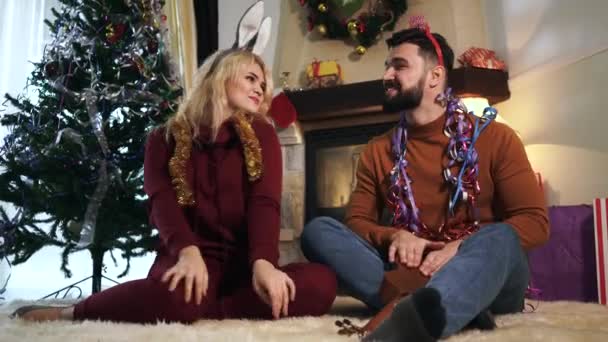 Vrolijke man en vrouw zitten voor de versierde kerstboom en dansen. Gelukkige blanke man en vrouw die thuis rusten op oudejaarsavond. Feestdagen, feesten. — Stockvideo