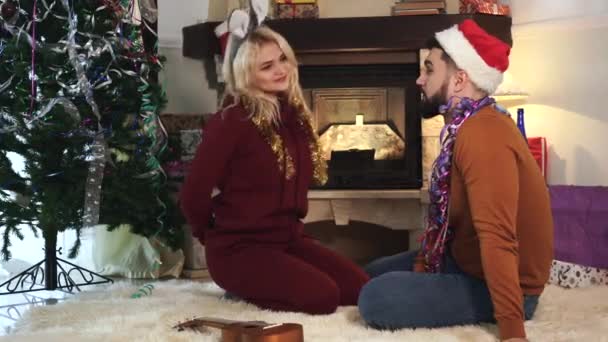 Positiv blond vit kvinna sitter med make framför öppen spis och julgran, hålla gåva bakom ryggen. Le man i rött nyår hatt gissa i vilken hand hans fru håller närvarande. — Stockvideo