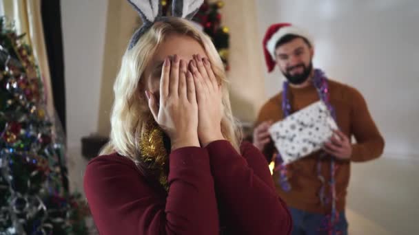 Gros plan de la jeune femme blanche blonde avec les yeux fermés au premier plan. Homme barbu déconcentré dans le chapeau de Noël tenant boîte cadeau à l'arrière-plan. Saint-Sylvestre, saison des fêtes . — Video