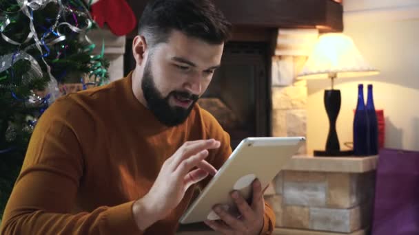 Ritratto di uomo bianco concentrato con barba nera seduto davanti all'albero di Natale e con tavoletta. Grave giovane ragazzo alla ricerca di regali online a Capodanno. Vacanze, sorpresa, regali . — Video Stock