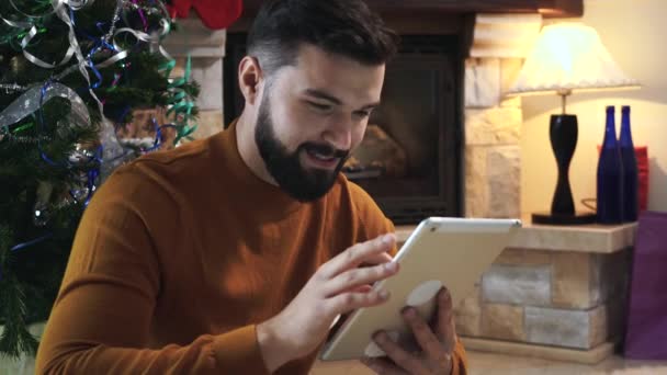 Κοντινό πλάνο του νεαρού Καυκάσιου που επιλέγει χριστουγεννιάτικο δώρο στο διαδίκτυο με τη χρήση tablet. Χαμογελώντας άνθρωπος κάνει το νέο έτος ψώνια μακριά στο σπίτι. Εποχή των διακοπών, ευτυχία, χαρά. — Αρχείο Βίντεο