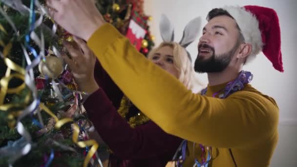 Gros plan d'un beau couple caucasien accrochant des décorations de Noël sur un arbre du Nouvel An et admirant leur travail. Jeune mari et femme décorant la maison la veille de Noël. Célébration, bonheur . — Video