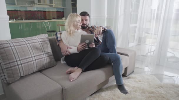 積極的な白人男性と女性のソファに座って、タブレットを使用しています。彼らの家のためのオンライン購入をする夫と妻。Eショッピング、無線技術、インターネット. — ストック動画