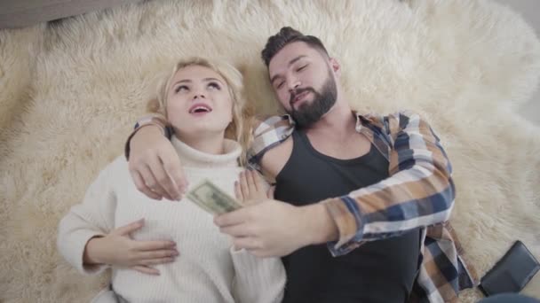 幸せな豊かな白人のカップルが柔らかいカーペットの上に横たわって、ドルを数える。白いセーターの金髪の女の子とハンサムなひげを生やした男が自宅で休んでいます。富、幸福、余暇. — ストック動画