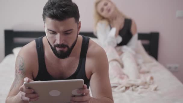 Portré felnőtt kaukázusi férfi ül az ágyon, és videojátékok segítségével tabletta. Csábító szőke nő a háttérben, aki a barátjára vagy a férjére vár. Játékok függőség, házasság problémák. — Stock videók