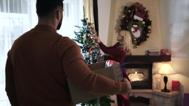 Camera volgende volwassen blanke man wandelen met doos naar mooie vrouw versieren kerstboom. Echtgenoot brengt decoraties voor zijn vrouw. Jong stel brengt oudejaarsavond samen thuis door. — Stockvideo