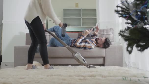 Неузнаваемая молодая кавказская женщина пылесосит ковер дома, когда её муж лежит на диване. Жена швыряла грязные носки на мужа и выходила из комнаты. Домашняя работа, поддержка, брак . — стоковое видео