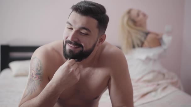 Close-up face of satisfied Caucasian man with tattoo on shoulder sitting on bed. Young blond woman at the background touching tenderly her hair and shoulders. — 비디오