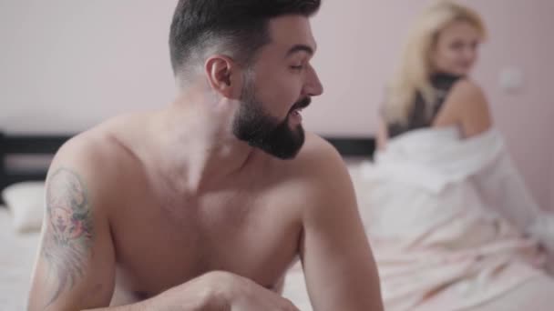 Porträtt av ung vit skäggig man tittar tillbaka på nöjd blond kvinna sitter på sängen i bakgrunden. Leende kille vilar i sovrummet efter sex. — Stockvideo