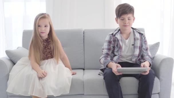 Portré két fehér gyerekről, akik a kanapén ülnek. A fiú néz valamit a tabletképernyőn, a lány gyönyörű ruhában elnéz. Első szerelem, szabadidő.. — Stock videók