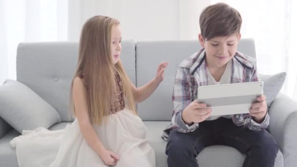两个白人孩子坐在沙发上。 可爱的白人女孩摸着男孩的肩膀挥手。 男孩做不满意的面部表情,回头看平板电脑屏幕. 飞舞，初恋. — 图库视频影像