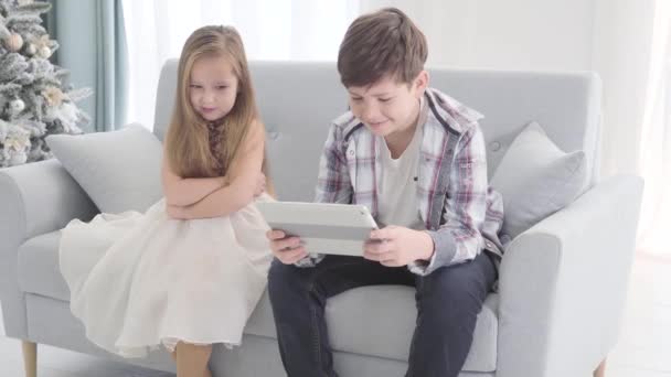 Retrato de niña caucásica ofendida sentada con las manos cruzadas junto a un chico sosteniendo la tableta. Tipo morena haciendo caso omiso de niño bonito en vestido hermoso. Infancia, primer amor, Nochevieja . — Vídeo de stock