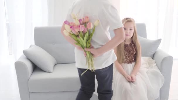 Vista posterior de chico caucásico en camiseta blanca y jeans llegando a chica bonita con un ramo de tulipanes. Niño sorprendido en hermoso vestido que cubre la boca con la mano. Primer amor, niños coqueteando . — Vídeos de Stock