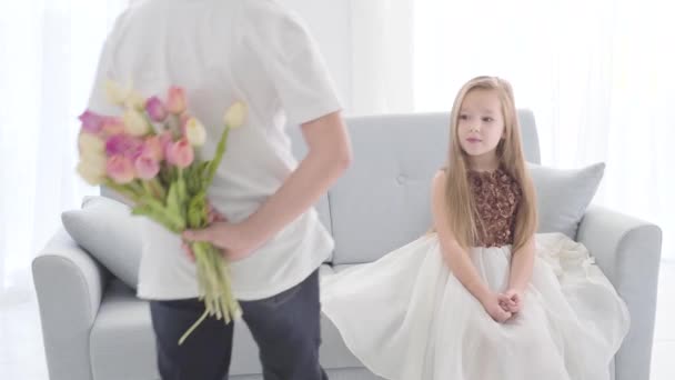 Unrecognizable Caucasian boy in casual clothes coming to cute girl with a bouquet of tulips behind his back. Surprised little lady in beautiful dress covering mouth with hand. First love, flirting. — Stock Video