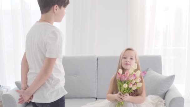 Jolie enfant caucasien en robe assise sur le canapé avec un bouquet de tulipes et regardant le garçon. Enfants amoureux, amoureux, innocents . — Video
