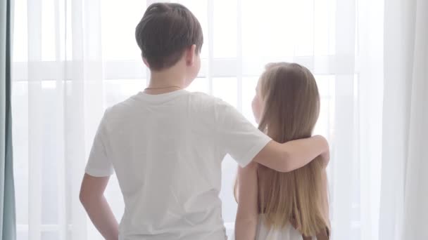 Back view of Caucasian boy and girl standing together and looking out the window. Brunette guy in white T-shirt holding hand on little ladys shoulder. First love, childhood. — Stock Video