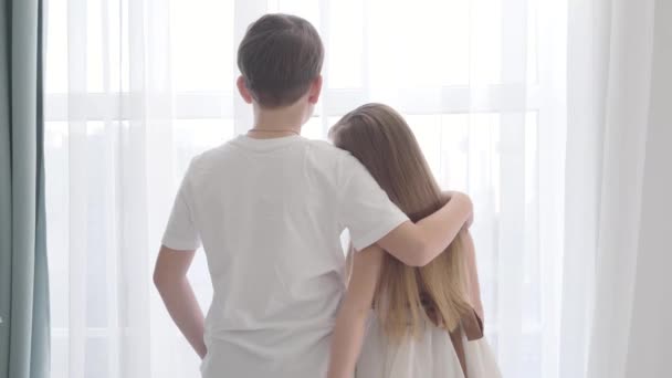 Kamera nähert sich kaukasischen Mädchen und Jungen, die zusammen vor dem Fenster stehen. Kinder, die sich umarmen und nach draußen schauen. erste Liebe, Glück, Gemütlichkeit. — Stockvideo