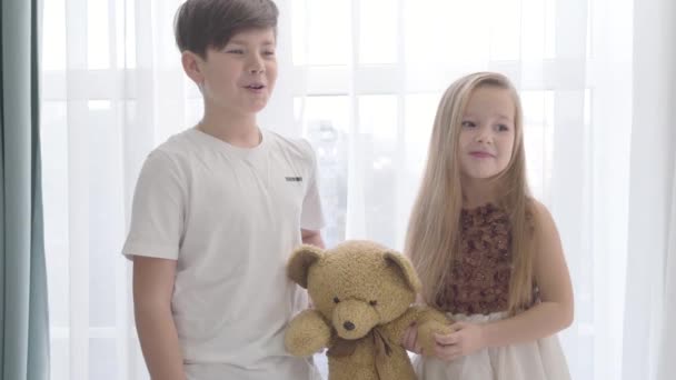 Retrato de niño y niña caucásicos sosteniendo oso de peluche. Chico alegre sonriendo y hablando, linda señorita en hermoso vestido mirando hacia otro lado. Infancia, primer amor, unión . — Vídeo de stock