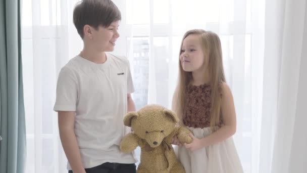 Petite fille et garçon caucasiens tenant un ours en peluche, se regardant et se souriant. Des enfants heureux passent du temps ensemble à l'intérieur. Enfance, premier amour, union . — Video