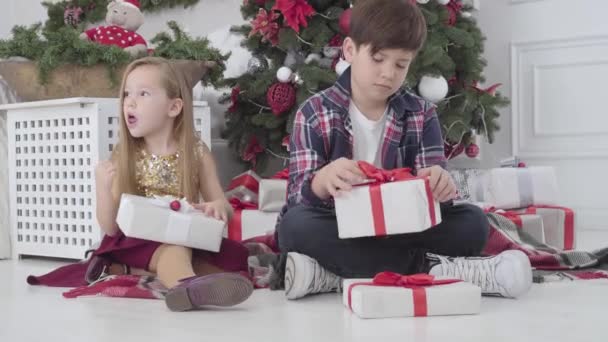 Portrait d'enfants caucasiens mignons assis sous le sapin de Noël avec des boîtes-cadeaux et regardant autour. Fille et garçon recevant des cadeaux la veille du Nouvel An à la maison. Vacances, loisirs . — Video