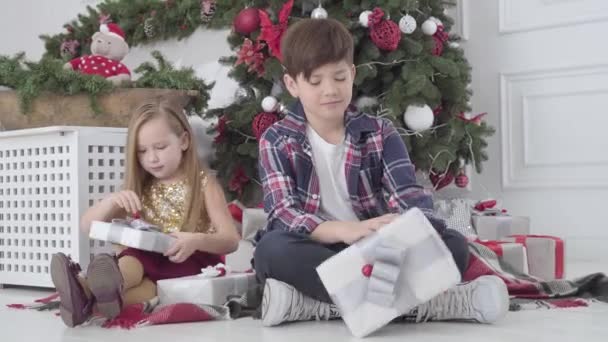 Kleine jongen en meisje met geschenken als zittend onder nieuwjaarsboom. Portret van gelukkige kinderen op kerstavond. Vakantie, feest, kindertijd. — Stockvideo