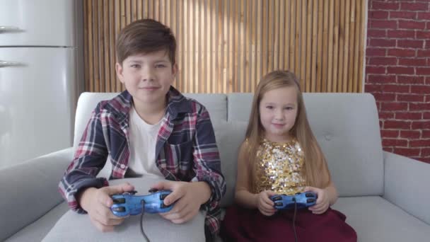 Двое белых детей играют в видеоигры, сидя дома на диване. Мальчик и девочка побеждают, делают победный жест и обнимаются. Отдых, детство, счастье . — стоковое видео
