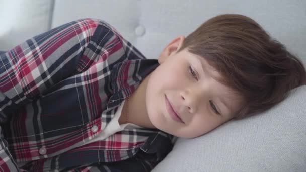 Close-up retrato de menino bonito caucasiano com cabelos castanhos e olhos cinzentos deitado em travesseiro macio. Criança sorridente descansando no sofá em casa nos fins de semana. Infância, lazer. estilo de vida, felicidade . — Vídeo de Stock