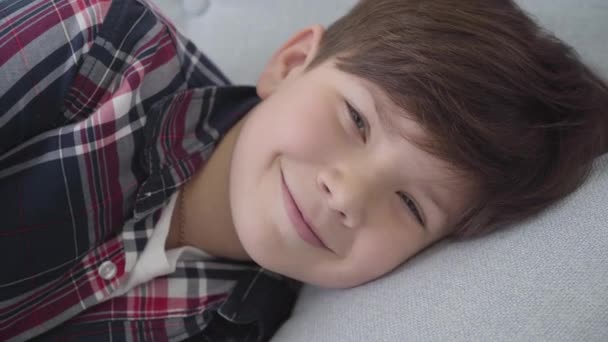 Primer plano del chico caucásico con el pelo castaño y los ojos grises mirando a la cámara y sonriendo. Niño feliz acostado en un sofá blando en casa. Infancia, ocio. estilo de vida, felicidad . — Vídeos de Stock