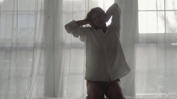 Porträtt av en söt vit kvinna som sträcker sig på sängen på morgonen. Blond tjej i vit skjorta och spetsunderkläder vaknar hemma. Lugn, lycka, sensualitet. — Stockvideo