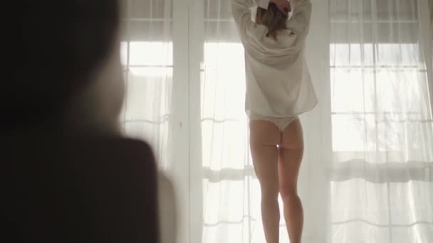 Back view of Caucasian girl in lace panties and white shirt standing in front of big window and stretching. Young attractive woman resting in bedroom in early morning. Leisure, lifestyle, silence. — Stock Video