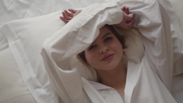 Portret van een jong blank meisje dat de ogen sluit met de handen en zich weer verstopt. Lachende blonde vrouw in wit shirt liggend op zacht bed. Lifestyle, geluk, ontspanning. — Stockvideo