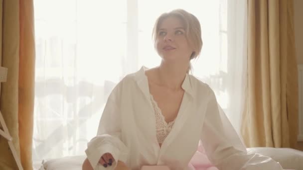 Porträt einer hübschen blonden Kaukasierin in Spitzenunterwäsche und weißem Hemd, die auf dem Bett sitzt und lächelt. attraktive junge Frau, die morgens im Sonnenlicht ruht. Ruhe, Lebensstil, Entspannung. — Stockvideo