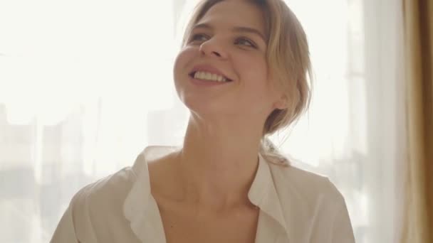 Close-up gezicht van lachende vrouw in wit shirt zittend in het zonlicht en glimlachend. Sensueel blank meisje dat 's morgens in de slaapkamer rust. Rust, levensstijl, ontspanning. — Stockvideo