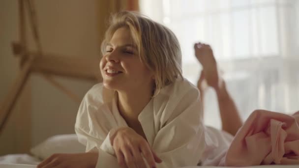 Porträtt av leende vit blond flicka liggande på mjuk säng och skakande ben. Glada kvinna i vit skjorta vilar i sitt sovrum hemma. Livsstil, lycka, fritid. — Stockvideo