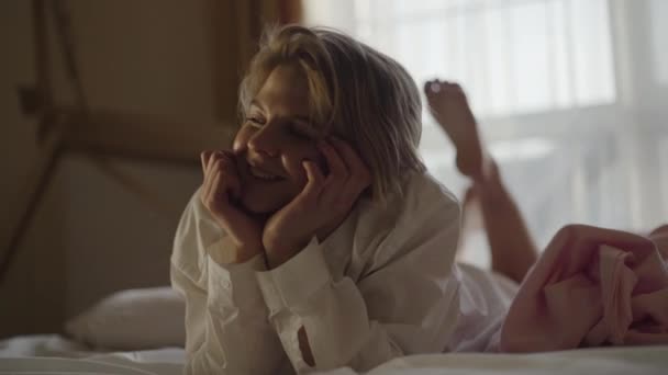 Portret van een blond blank meisje dat op een zacht bed ligt met handen en trillende benen. Positieve jonge vrouw in wit shirt rustend in haar slaapkamer thuis. Lifestyle, geluk, vrije tijd. — Stockvideo