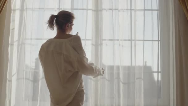 Αισθησιακή νεαρή λευκή γυναίκα με λευκό πουκάμισο έρχεται στο μεγάλο παράθυρο το πρωί. Πορτρέτο της ελκυστικό κορίτσι στέκεται σε ακτίνες του ήλιου και συγκινητικές κουρτίνες. Χαλάρωση, αναψυχή, τρόπος ζωής. — Αρχείο Βίντεο
