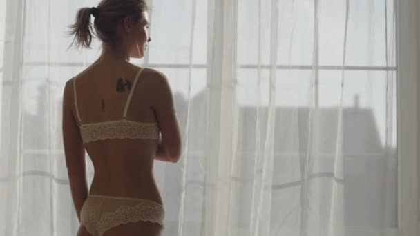 Achteraanzicht portret van een aantrekkelijk blank meisje dat in het zonlicht voor het raam staat. Sensuele vrouw in kanten ondergoed die binnen rust. Sensualiteit, levensstijl. — Stockvideo