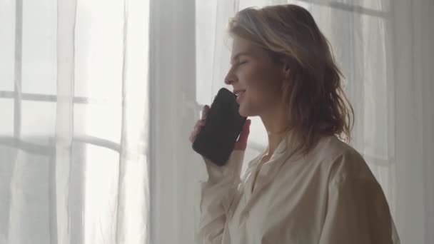 Vista laterale ritratto di giovane donna bionda carina in piedi sotto i raggi del sole davanti alla finestra e parlando al telefono. Giovane donna caucasica che tiene smartphone e sorride . — Video Stock