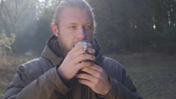 Close-up portrait of bearded Caucasian man with grey eyes standing in the forest and drinking hot tea from steel mug. Handsome guy resting outdoors alone. Lifestyle, relaxation, leisure. — 비디오