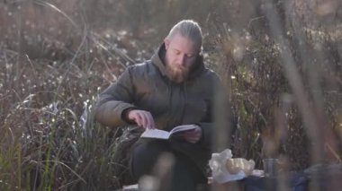 Sonbahar ormanında ağaç gövdesinde oturup kitap okuyan düşünceli beyaz adamın portresi. Erkek turist açık havada tatilin tadını çıkarıyor. Boş zaman, yaşam tarzı, hobi.
