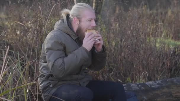 Side view of adult Caucasian man sitting on tree trunk and eating hot-dog. Confident male tourist resting outdoors alone. Lifestyle, relaxation, leisure. — Stock Video