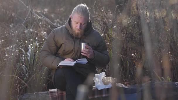 Thoughtful Caucasian man sitting on tree trunk, reading and drinking hot tea from steel mug. Lonely male tourist enjoying resting outdoors. Leisure, lifestyle, hobby. — 비디오
