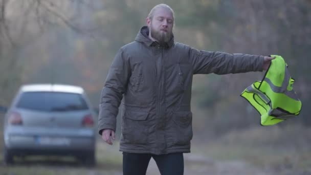 Portret van een ongelukkige blanke toerist die op bosweg staat en een groen vest schudt. Een man voor zijn kapotte auto gooit wanhopig vest weg. Liften, toerisme, hobby. — Stockvideo