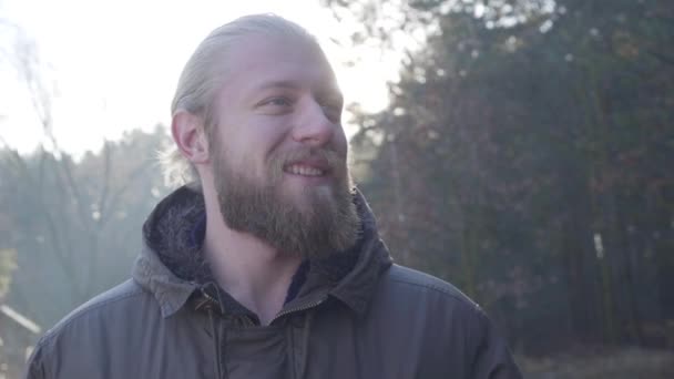 Close-up de belo homem caucasiano com olhos cinzentos em pé à luz do sol na floresta de outono. Turista sorridente desfrutando de seus fins de semana ao ar livre. Estilo de vida, viagens, turismo . — Vídeo de Stock